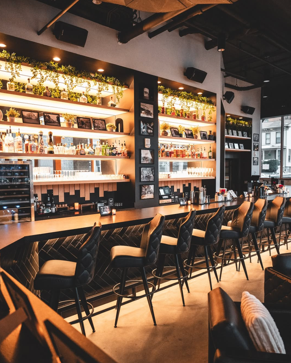 The long bar at 1824 lined with leather stools and backlit shelves of spirits and greenery